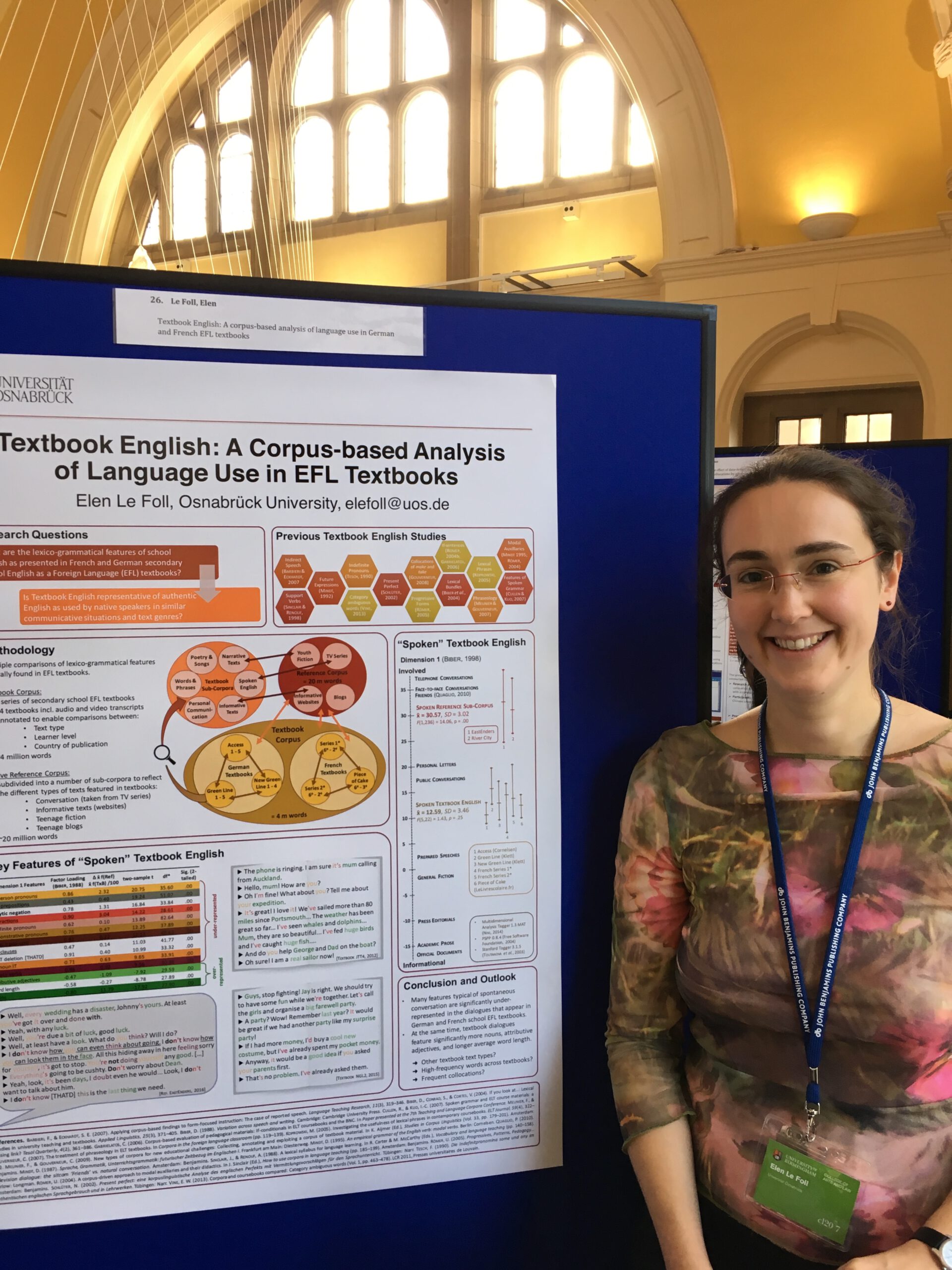 Photo of a white woman with glasses in her 30s smiling, wearing a floral top and a conference badge. She is standing in front of an academic poster entitled: Textbook English: A Corpus-Based Analysis of Language Use in EFL Textbooks.